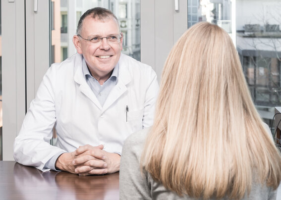 Der Neurologe, Dr. Joachim Hartmann sitzt an seinem Schreibtisch und spricht mit einer Patientin.